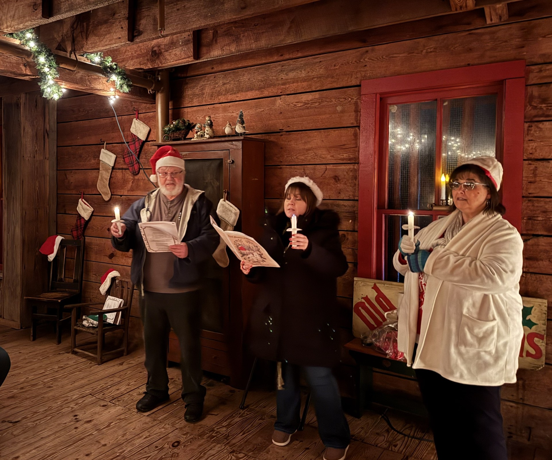 A family of three, father, mother and daughter lead holiday carols in a dimly lit wooden structure. All three are holding burning candles, santa hats and have printed song books in their hands singing.