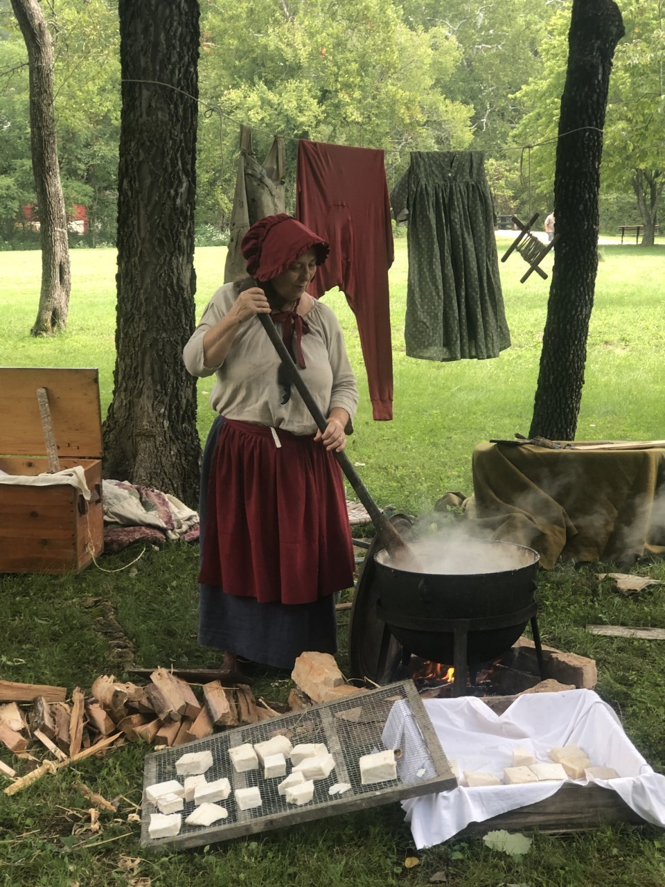A lady dressed in old fashioned clothing next to a large iron pot over the fire making lye soap. The white soap can be seen in the pot. She is stirring it with a large wooden paddle.