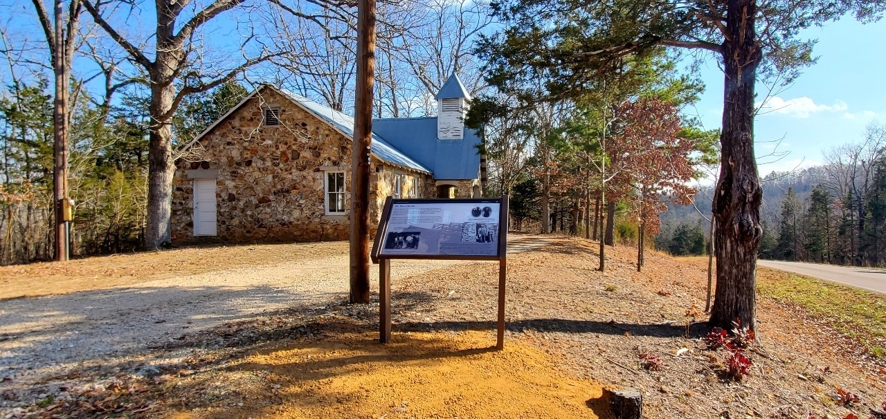 A rock stone structured church in the distance with a wayside in front of it. Highway to the right and surrounded by trees.