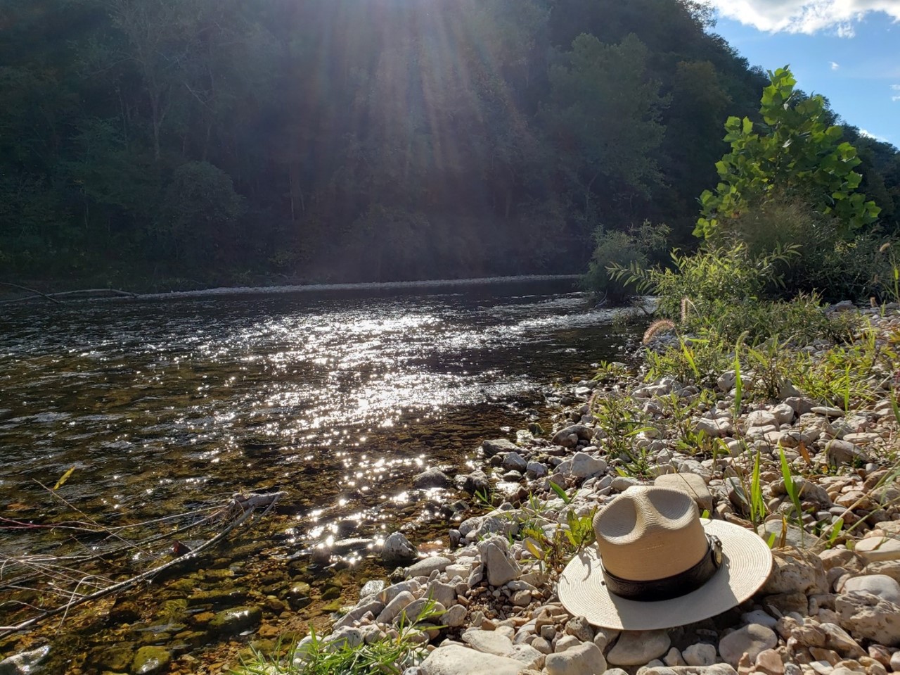 A clear flowing river with sun reflecting off of it, a flat ranger hat on a gravel bar on the edge of the river