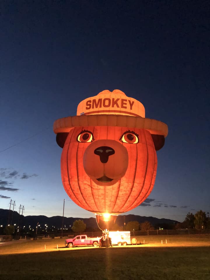 Smokey Bear hot air balloon lighted up inside and glowing during the night with a crew and support vehicles below it.
