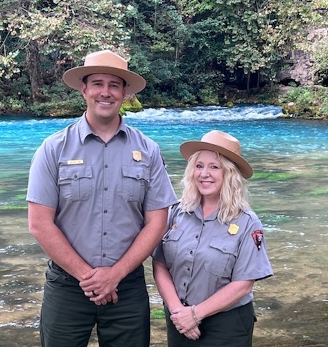 thumbnail_Stacey William 1a Two park rangers in flat hats and uniforms stand next to a blue and clear large spring. They are smiling at the camera.