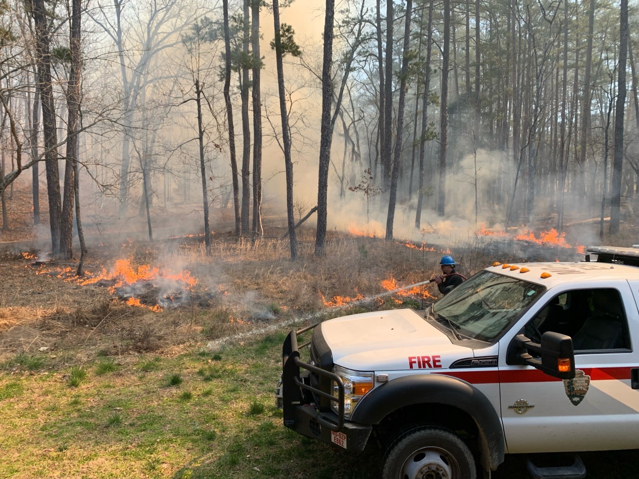 A wildland fire fighter using a hose to spray an area of green grass next to dry grass that is active with fire. A national park service fire truck is parked next to the firefighter.