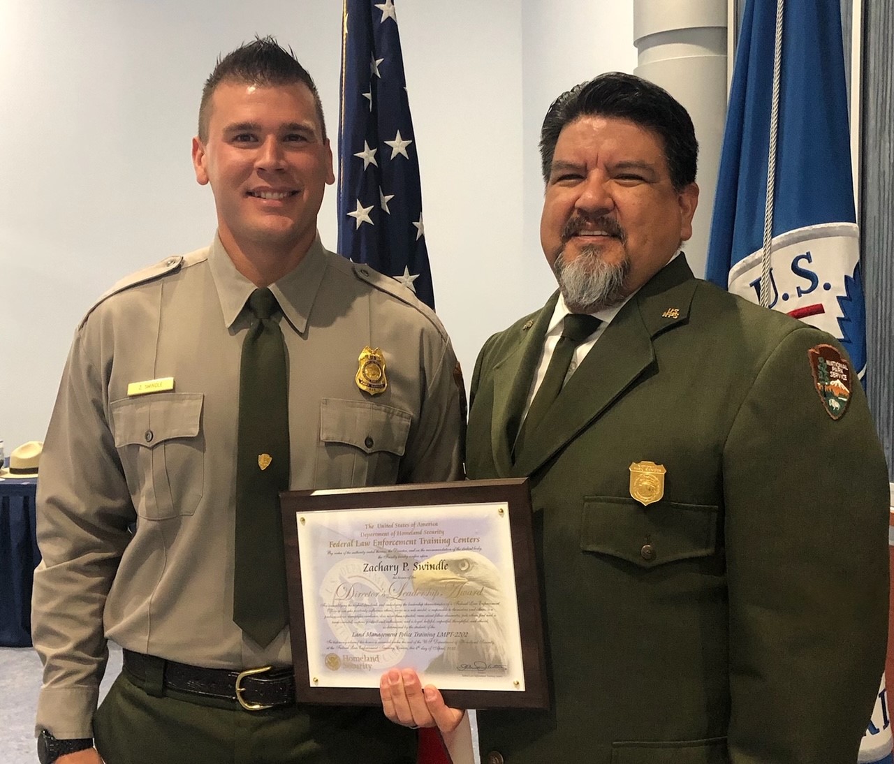 thumbnail_Zack Swindle and Chuck Sams Two men stand together holding a square plaque between them in National Park Service green and grey attire. The U.S. Flag is behind them.