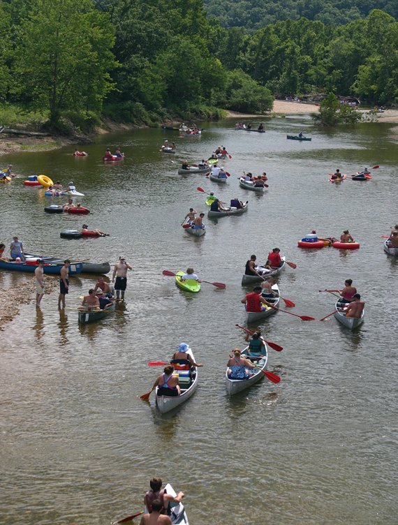 thumbnail_image001 Looking down a crystal clear river surrounded by forest. Several kayaks, canoes and tube with floaters in them during a summer day.