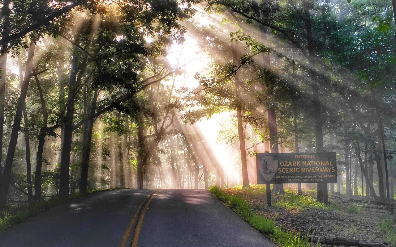 A color photo showing a asphalt road surrounded by a forest and brown Ozark National Scenic Riverways entrance sign beside the road. The suns rays are peeking through the forest canopy.
