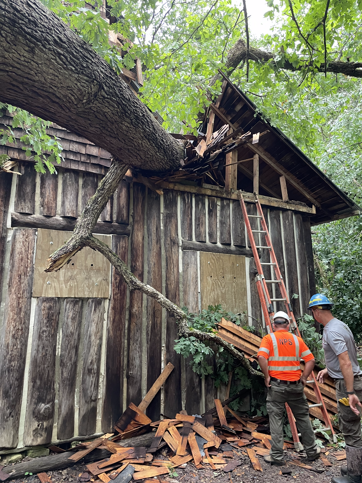 thumbnail_pulltite cabin-roof damage-01-reduced Two NPS employees with safety vest and hard hats. Several wooden shingles are laying beside the buidling. A large tree is leaning on the peak roof and has damaged the roof.