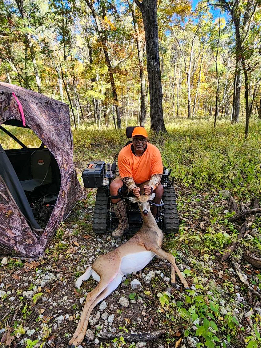 A man in a blaze orange hat and shirt sitting in a track chair in an open woodland setting displays a recently harvested whitetail deer.