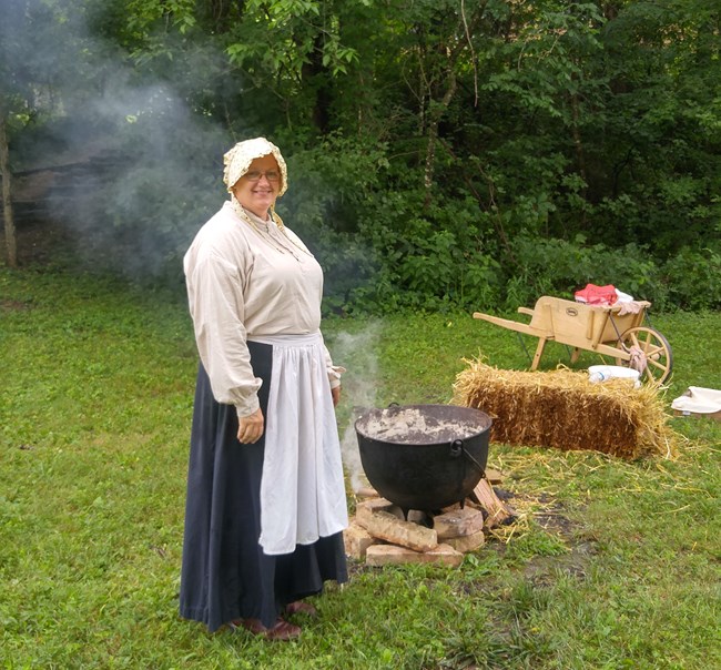 lady in pioneer dress and bonnet stirs lye soap in kettle