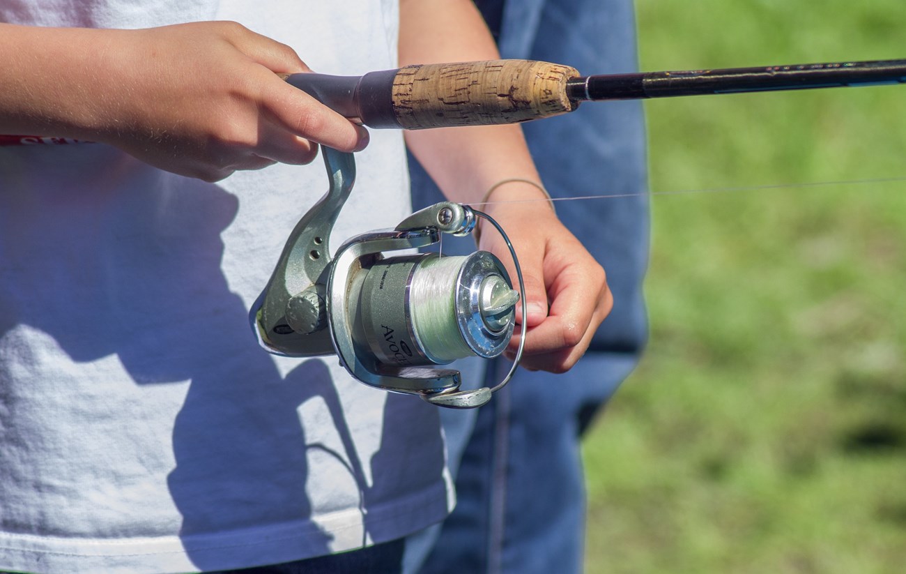 Photo of waist only of a person holding a spinning fishing rod in right hand and reeling with other, person is wearing a white shirt. A shadow from the fishing rod and reel are cast on their white shirt. Fishing reel is silver with white line.
