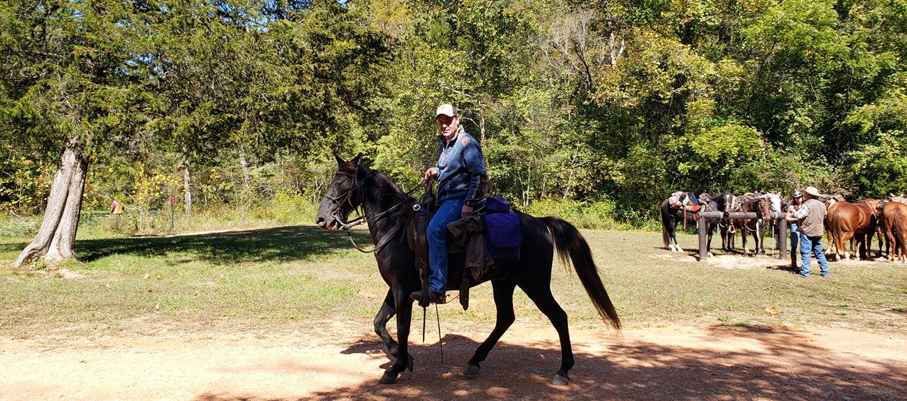 A man rides a horse along a gravel road, with hitching posts in the background.