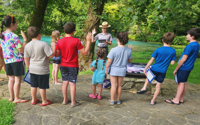 A group of children of various ages in summer clothes with right hands raised stand around a uniformed park ranger with her right hand raised and holding a junior ranger activity book in the other. In the background is a body of aqua blue water.