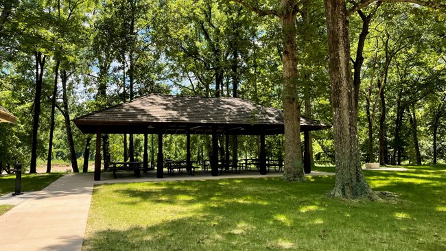 A picnic pavilion with concrete floor and hipped, wood shingle roof supported by 8 square metal posts stands in a forest clearing. A cut stone fire ring can be seen in the distance on the right side, and concrete sidewalks lead t and surround the pavilion