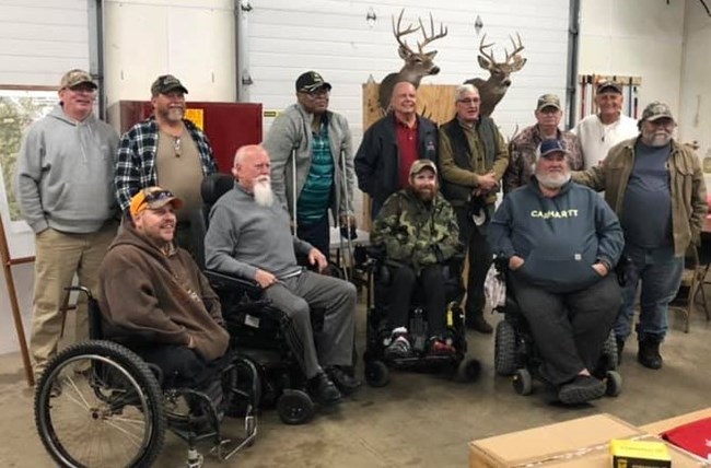 A group of hunters some standing, some in wheelchairs inside a building during a previous hunt gathering.