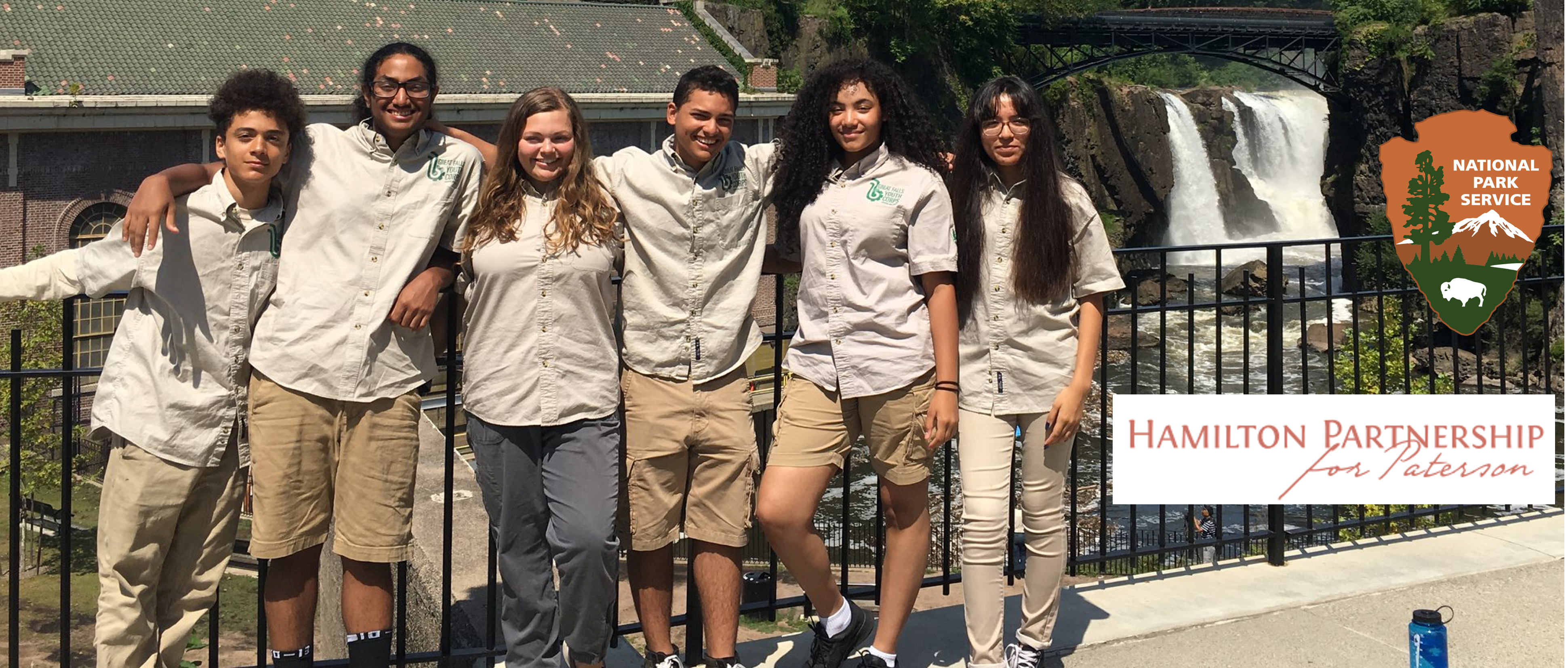 Six high school students in a straight line standing in front of a black fence with the Great Falls visible in the background on the right.  National Park Service and Hamilton Partnership for Paterson logos on far right.