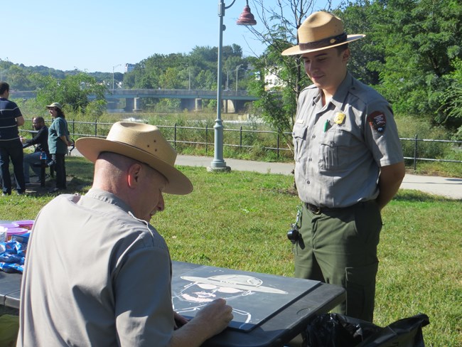A grey & green uniformed park ranger in a flat hat smiles as another at a table draws himself in chalk on a board