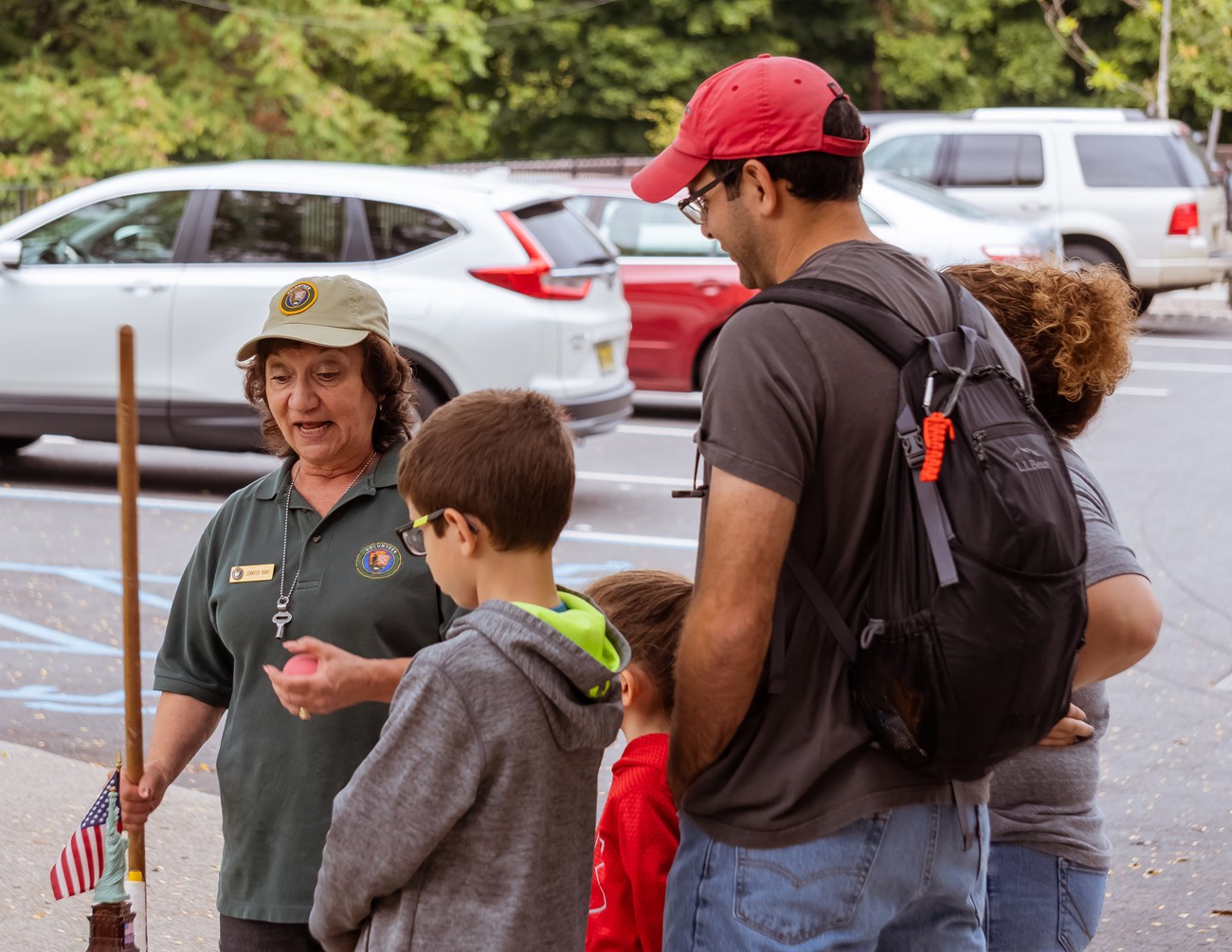 A green shirted volunteer speaks with a family as she shows them a collection of props including an American flag, a ball, & a model of the Statue of Liberty