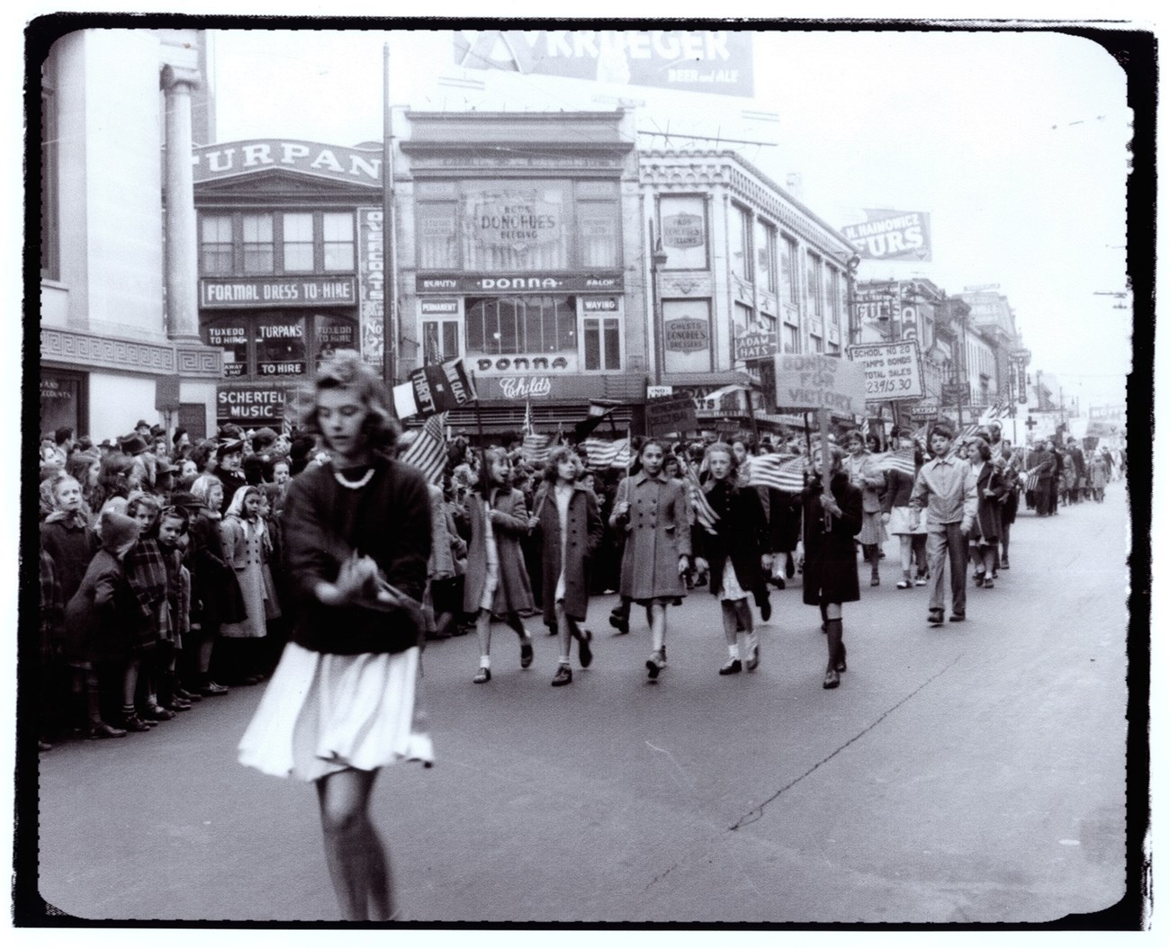 Black & White photo of youths marching in a 1943 Paterson NJ WWII Bond Drive. Carrying flags & signs, they pass crowds lining sidewalks lined with 3 & 4 story commercial buildings