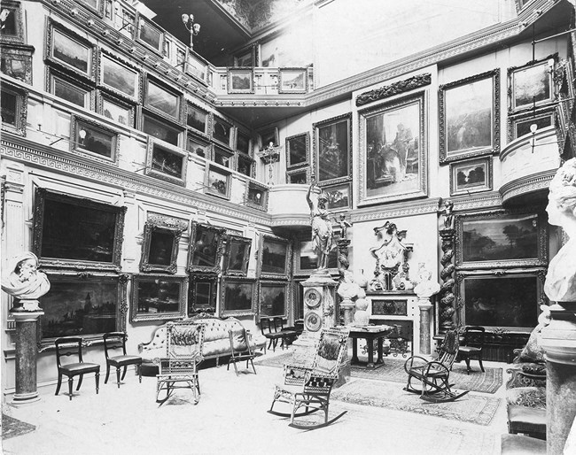 Black & white photo of a three-story room with overlooking balconies. The walls are crammed with massive paintings in ornate frames, & upholstered furniture crowd statues & busts on pedestals grouped around an ornate fireplace