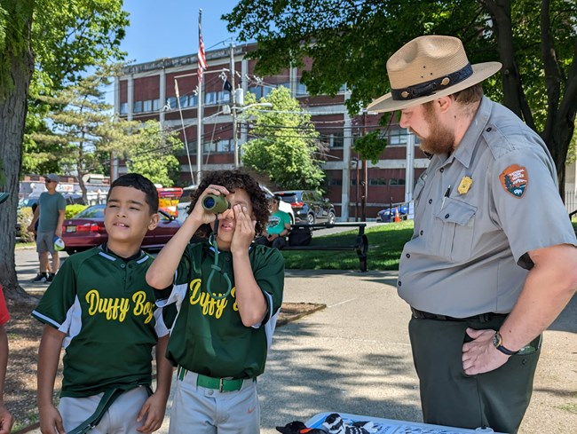 A park ranger stands at a table w/ plush birds beside two young boys looking forward - one holds a monocular to their eye and covers the other to see better