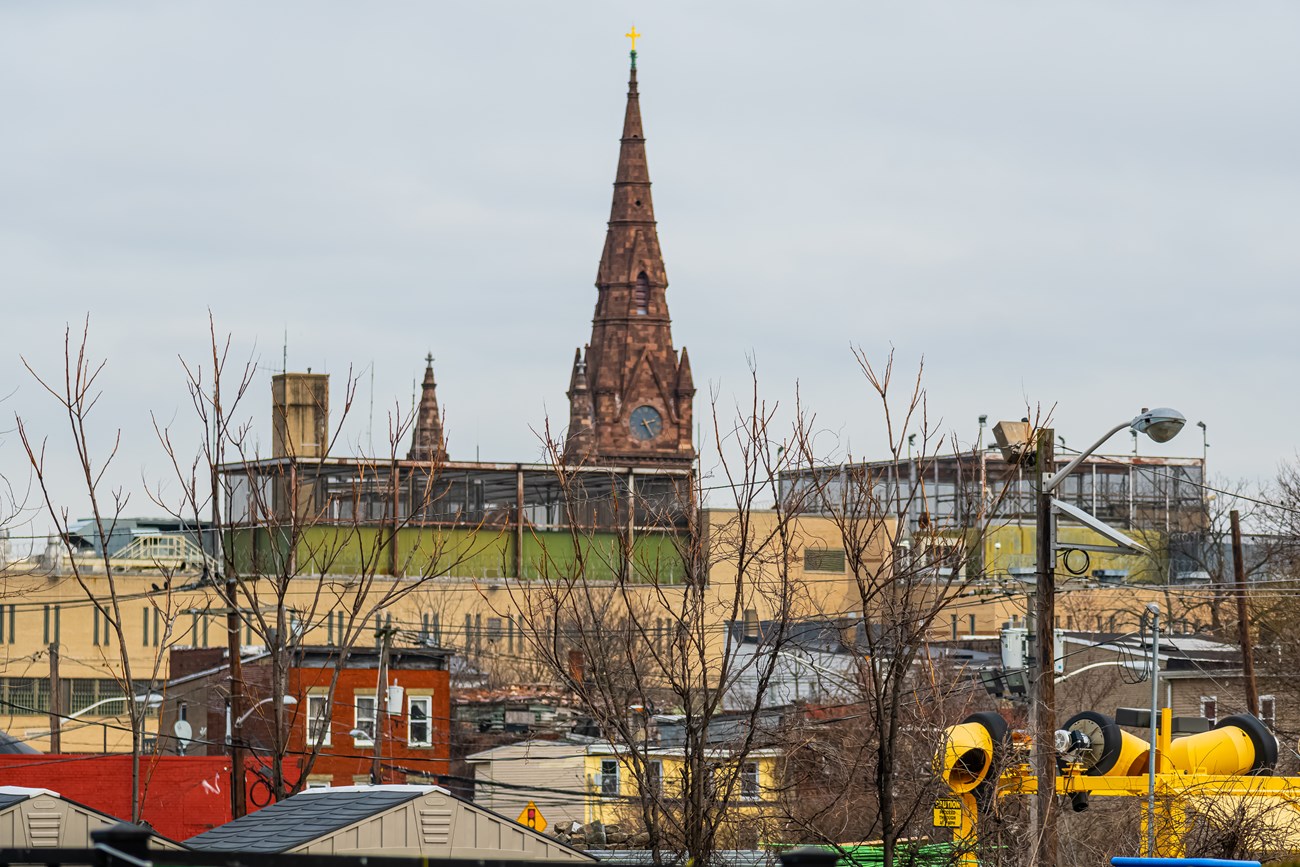 A wide shot of buildings & a neighborhood in Paterson, the brownish-red spire & clock tower of the Gothic-Revival Cathedral of St. John the Baptist rising in the background