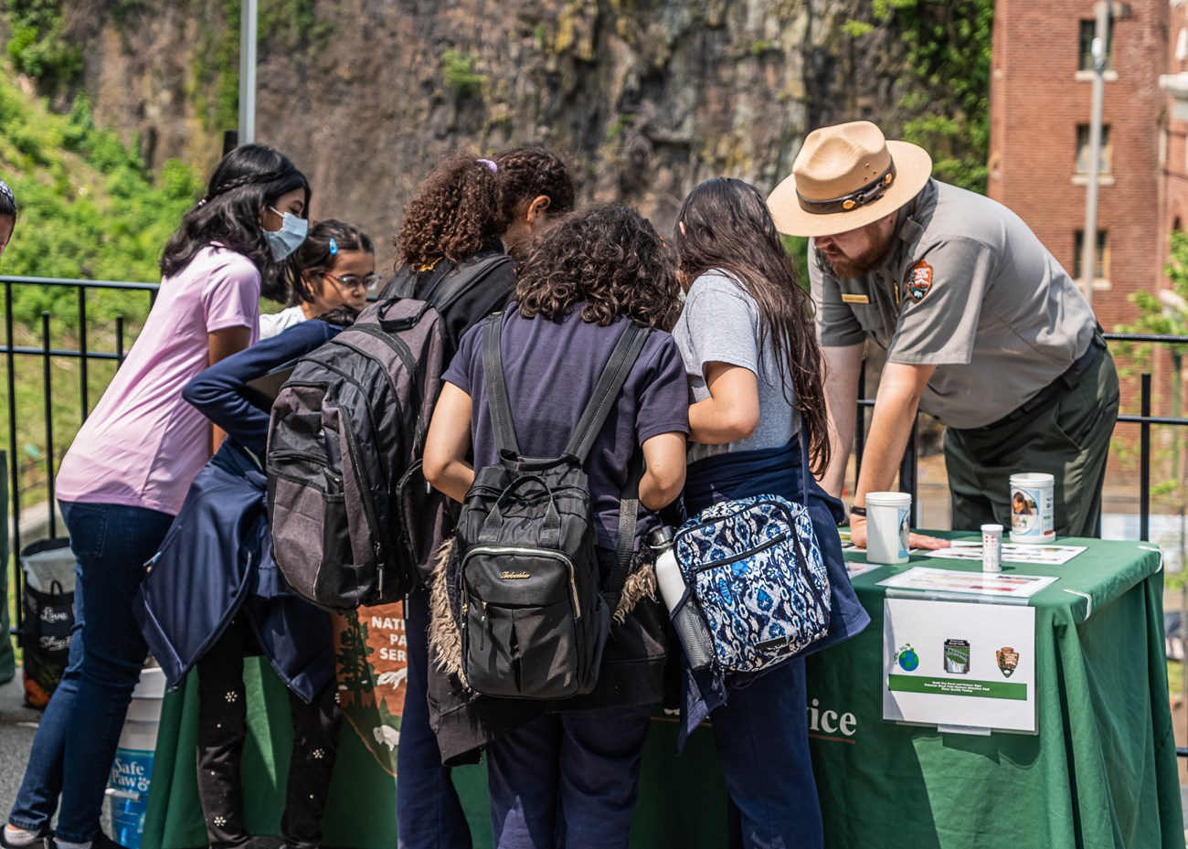 A park ranger leans over an activity table, working with a crowd of young students A park ranger leans over an activity table, working with a crowd of young students