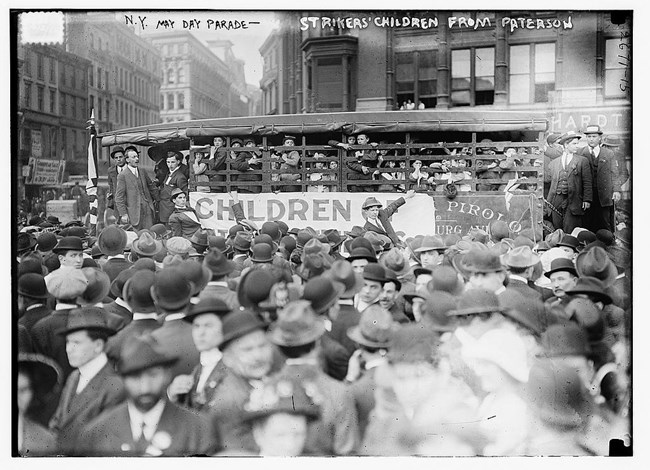 1913 black & white photo of a 1913 May Day parade shows a bus of striker's children from Paterson surrounded by a crowd of mostly men in bowler hats in downtown New York City