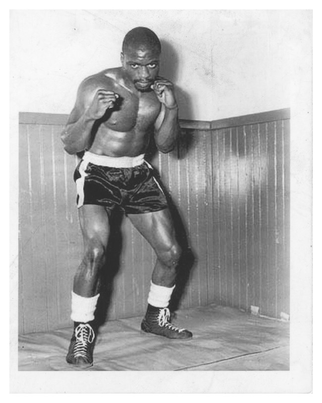 Black & white photo of Rubin "Hurricane" Carter. The black boxing athlete stands, arms raised, shirtless in boxing shorts and boots in a prepared stance
