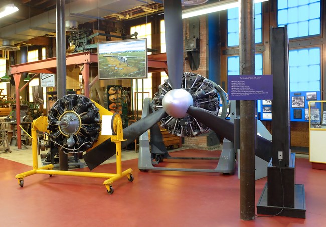 Two large radial aircraft engines, one on a yellow stand & one mounted with three massive propellors on a blue/grey stand, sit in a museum exhibit