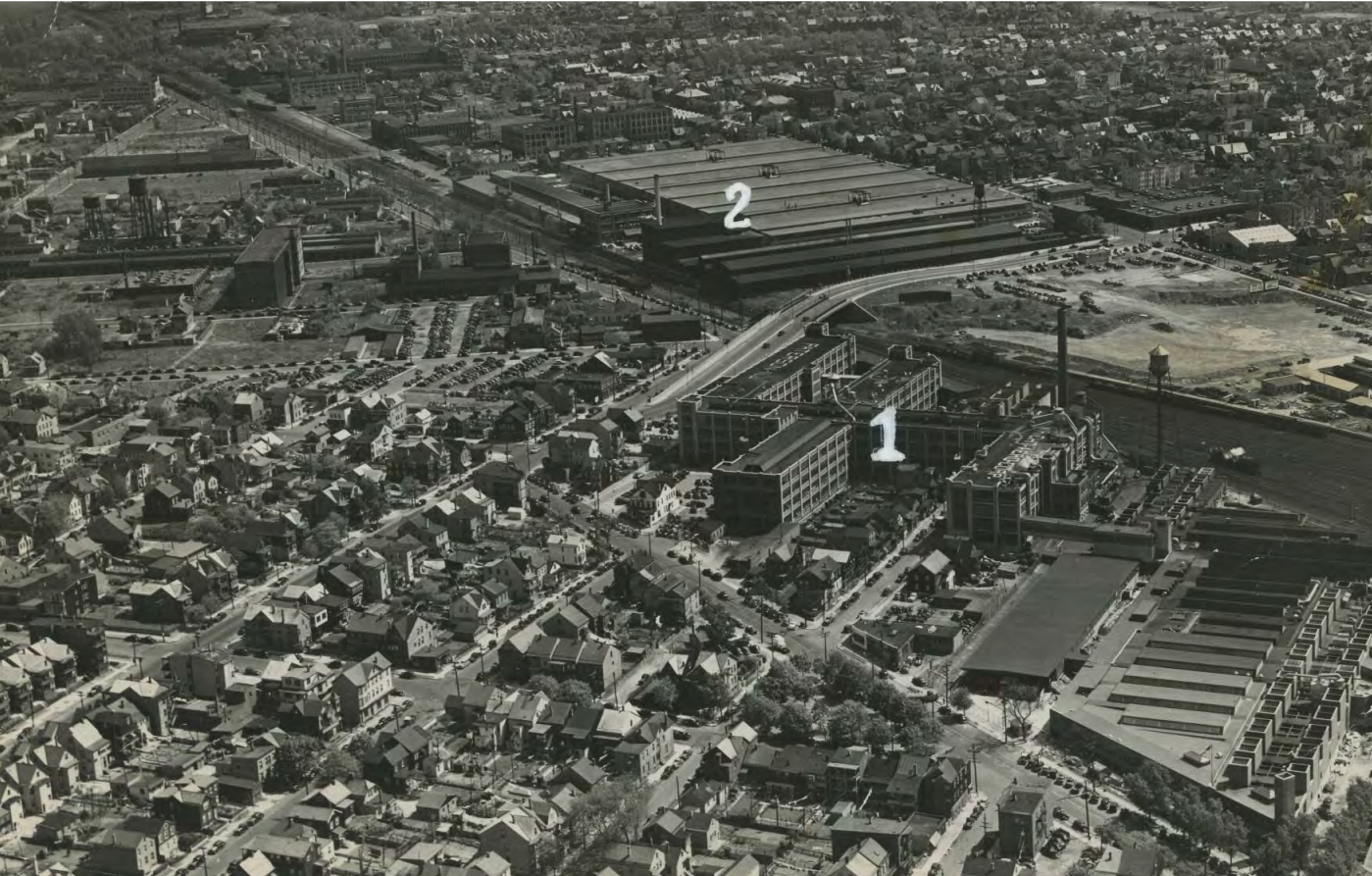 A 1940s aerial image of dense neighborhoods surrounding two massive manufacturing plants along a wide railroad line, numbered "1" & "2" for identification
