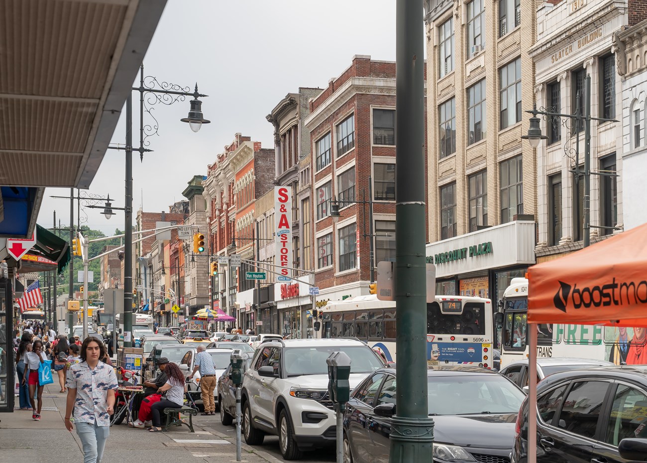 A busy downtown Paterson NJ street, crowded road lined with shops, restaurants, & pedestrians