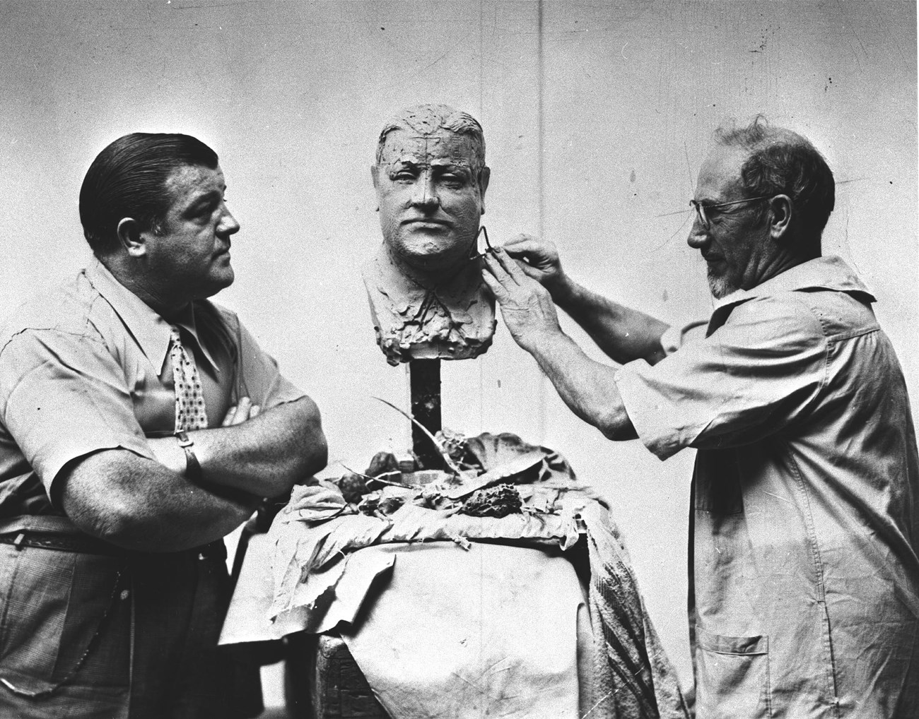 Black & white photo of sculptor Gaetano Federici, a balding man with a short white beard & glasses, carving a clay bust of Lou Costello as the comedian observes
