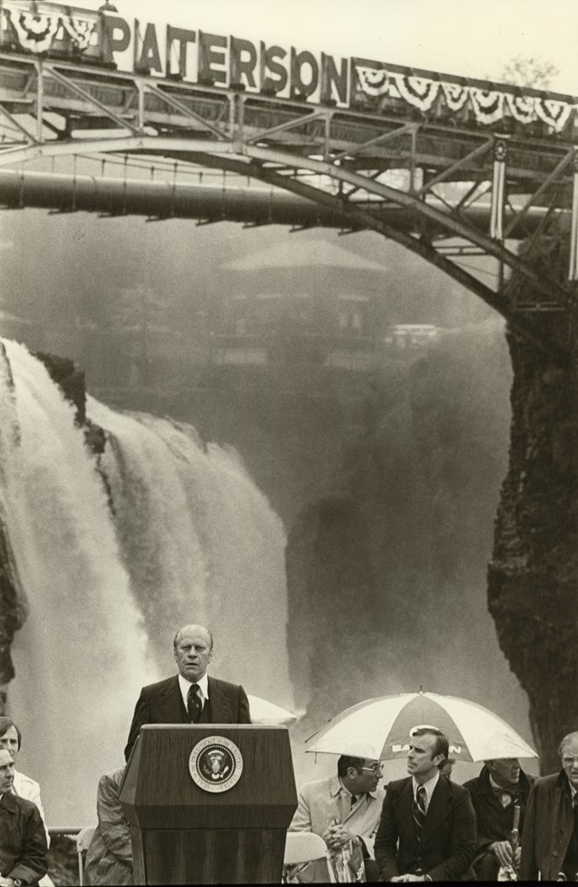 President Gerald Ford stands at a podium with the presidential seal beside seated dignitaries, the 77 ft. great falls roaring behind him framed overhead by an arched black metal bridge decked in patriotic bunting & a banner reading "Paterson"
