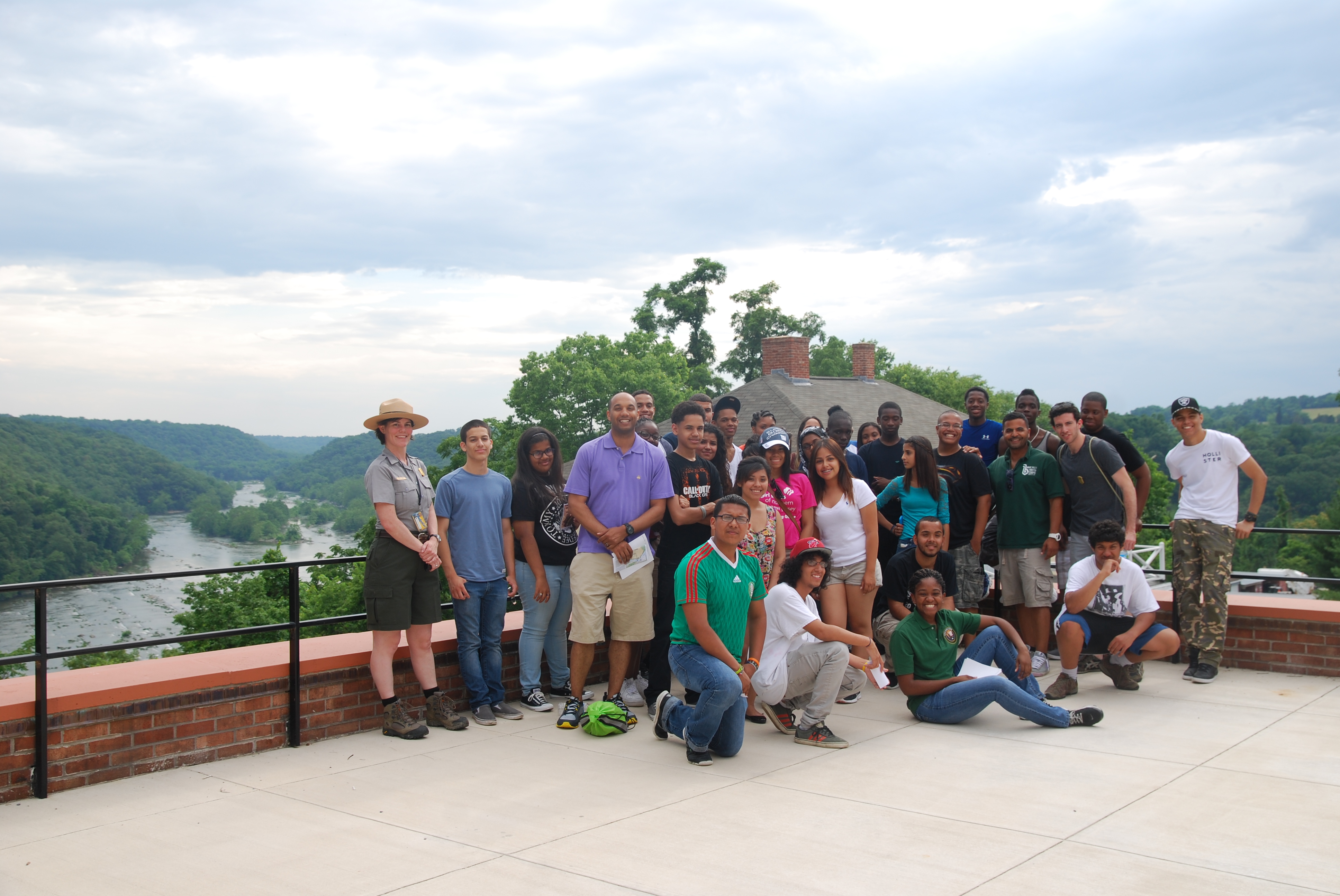 A group of high school youth corps members are standing with a park ranger in front of a river, green trees, and blue sky. Great Falls Youth Corps Visit Harpers Ferry