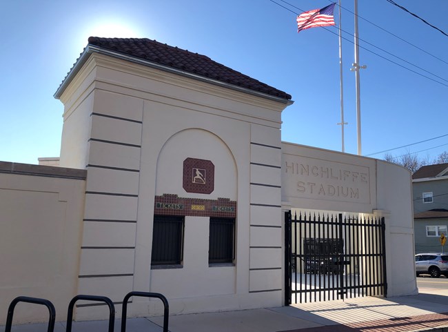 A white art-deco entrance to Hinchliffe Stadium, decorated with an ornamental gate, red terra-cotta roof tiles, and mosaics decorating the ticket booth. An American flag on a tall pole flies overhead