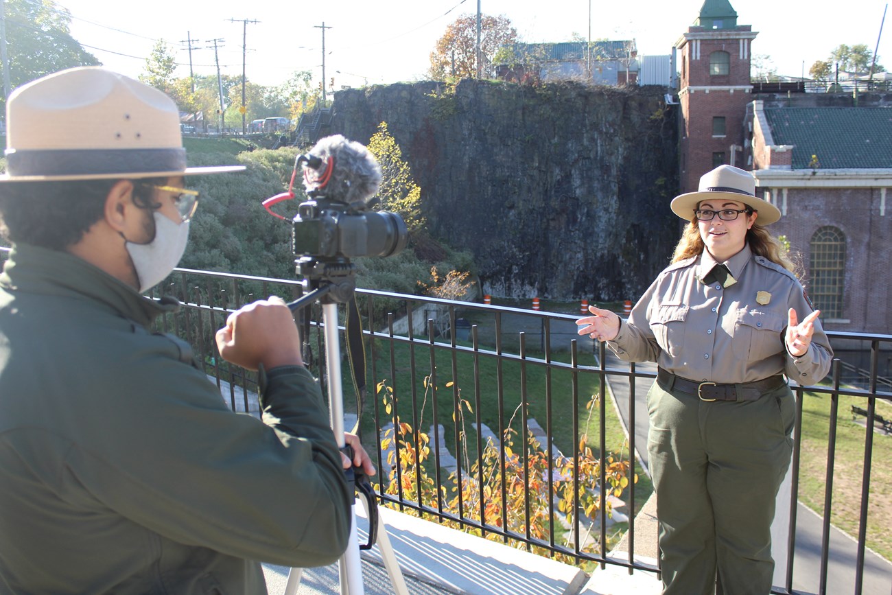 A park ranger films his colleague as she gives a virtual talk at a railing above a lower outdoor amphitheater & a brick hydroelectric power plant