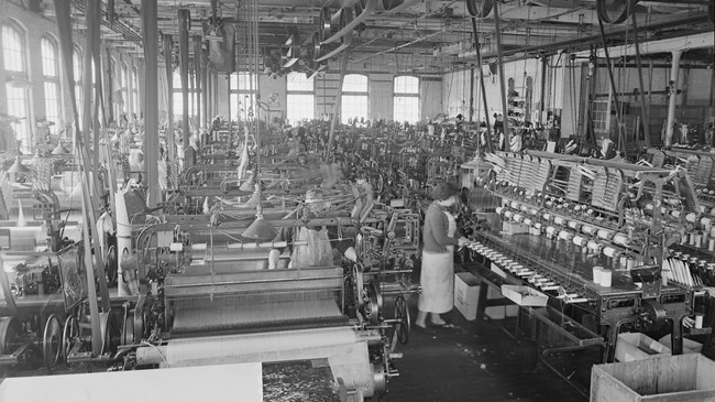 Black & white photo of workers in a silk mill c. 1941-42; overhead line shafts & belts drive looms & equipment in the large room well-lit by rows of large windows