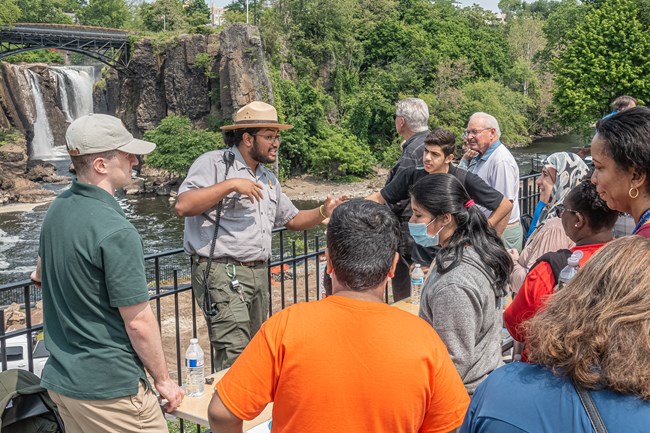A park ranger speaks with a crowd of visitors at a fence overlooking a 77 ft. waterfall framed by dark basalt cliffs & a black arched metal bridge