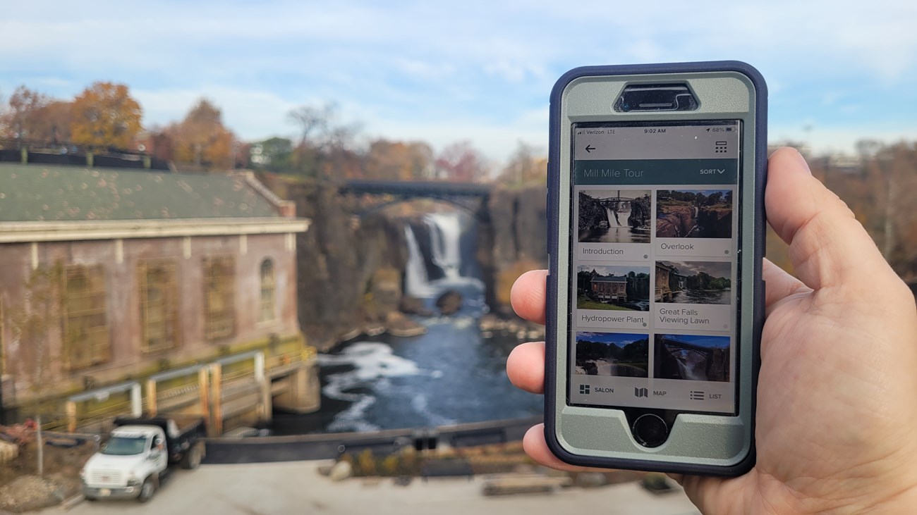 A hand holds a smartphone in view, displaying a guided audio app of Paterson Great Falls. In the background, a waterfall cascades 77 ft. between dark basalt cliffs, framed by an arched metal bridge & flanked by a brick hydroelectric station