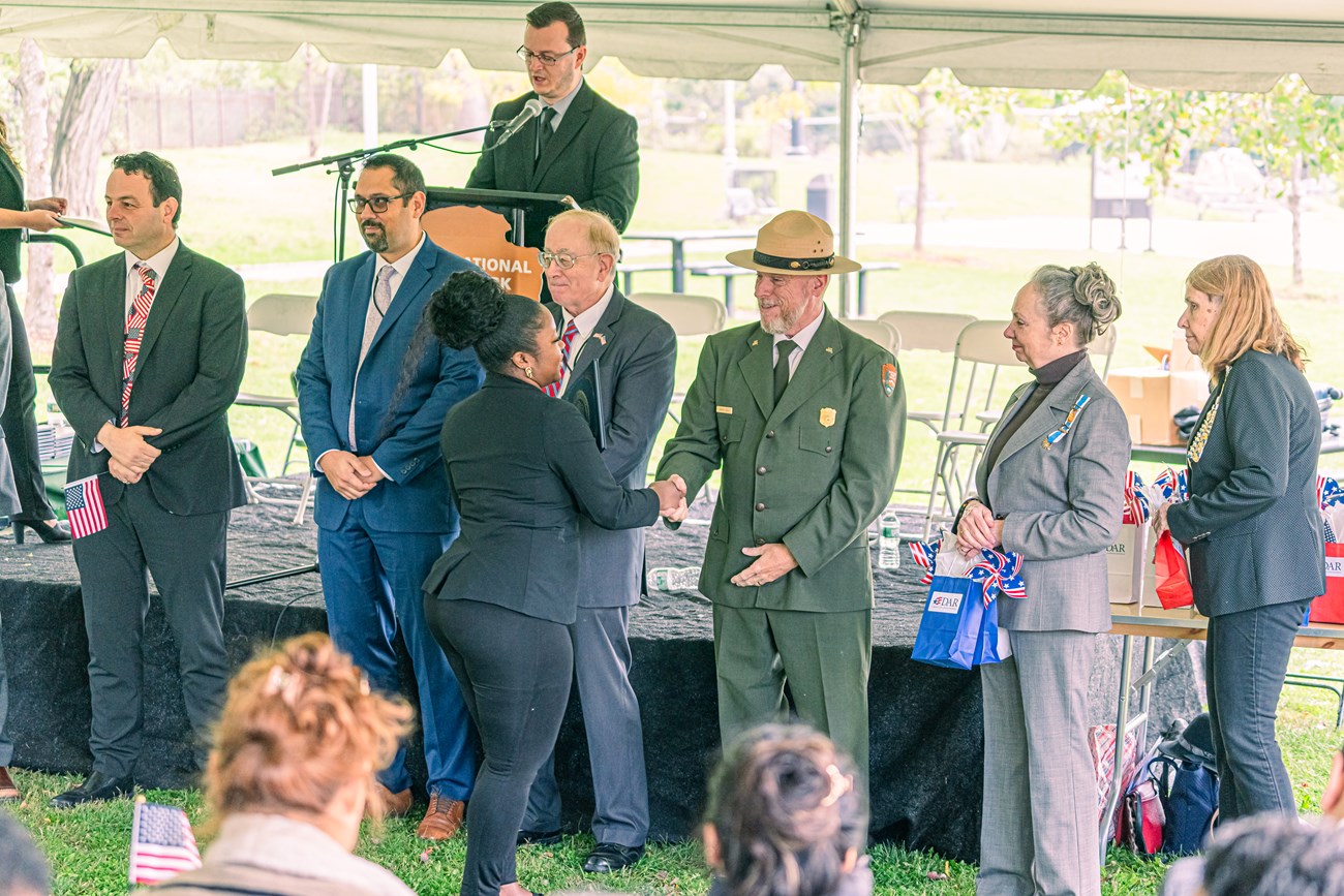 A park ranger in dress uniform shakes hands with a woman as other people in suits and dresses stand in a line beside him & on a podium