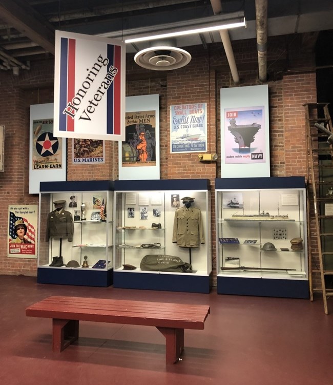 A red, white, & blue banner labeled "Honoring Veterans" hangs above wartime posters & display cases of uniforms, personal belongings, & military artifacts in a museum