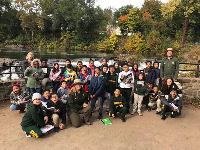 Park rangers stand & kneel beside a group of young school children standing at a waterfall overlook