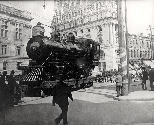 Black and white photo taken 9/21/1900 of a new 4-6-0 steam locomotive #265 on a heavy railed flatbed dragged by a team of 32 horses past tall downtown buildings in Paterson NJ as crowds look on