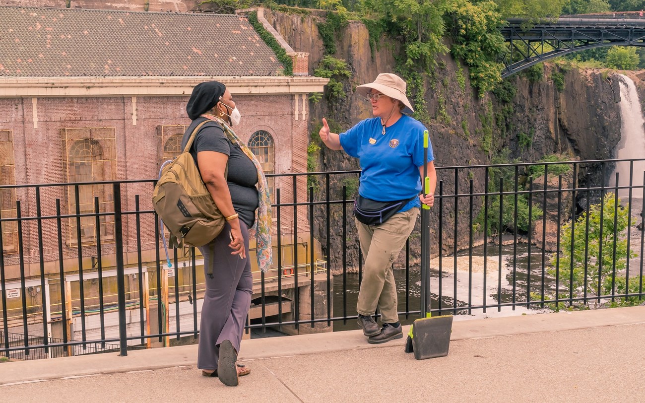 A volunteer in blue stands with a dustbin, talking to a woman with a waterfall, arched metal bridge, & brick hydroelectric plant in the background
