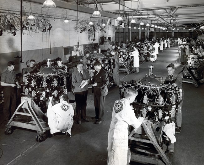 Black & white photo of workers in white technician's coats labeled "Wright Aero Corps" & dark overalls fit parts on an assembly line to rows of radial aircraft engines on rolling carts in a long, well-lit building as two managers confer