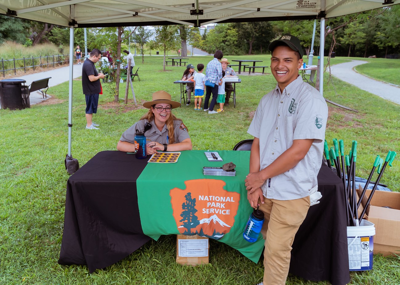 A seated park ranger in grey & green uniform with flat hat smiles at a table under a tent as a young man in a Great Falls Youth Corps shirt & hat smiles beside her