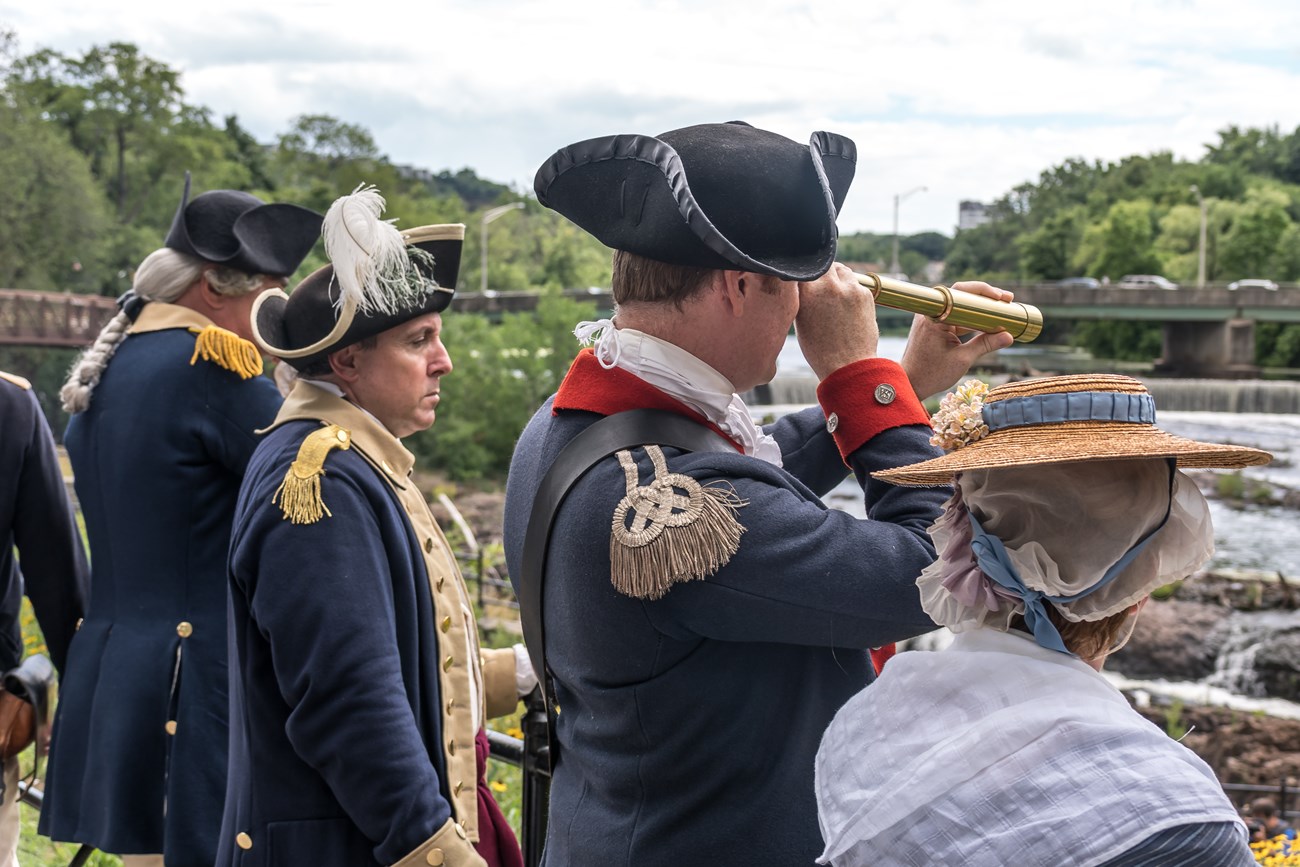 Men and women in American Revolutionary War uniforms & dress observe the Passaic River - one holds a brass telescope to their eye