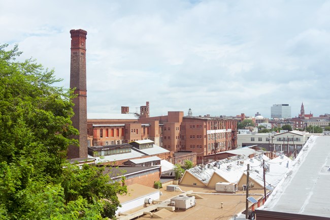 Tall, long brick mills with rows of windows and worn chimneys stand over smaller industrial buildings, office and civic buildings of Paterson NJ's skyline in the background