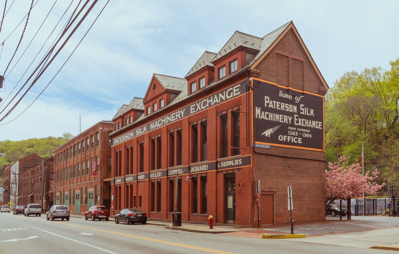 Red brick mills line a street; painted on the sides, the front one advertises the "Paterson Silk Machinery Exchange" & "Looms, warpers, winders, quillers, coppers, jacquards, & supplies."