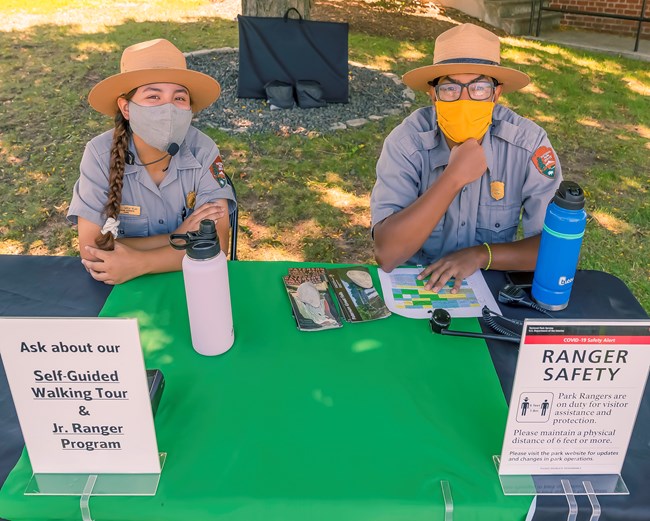 Park rangers at an outdoor table wear face masks - information stands list walking tours & Covid19 safety precautions